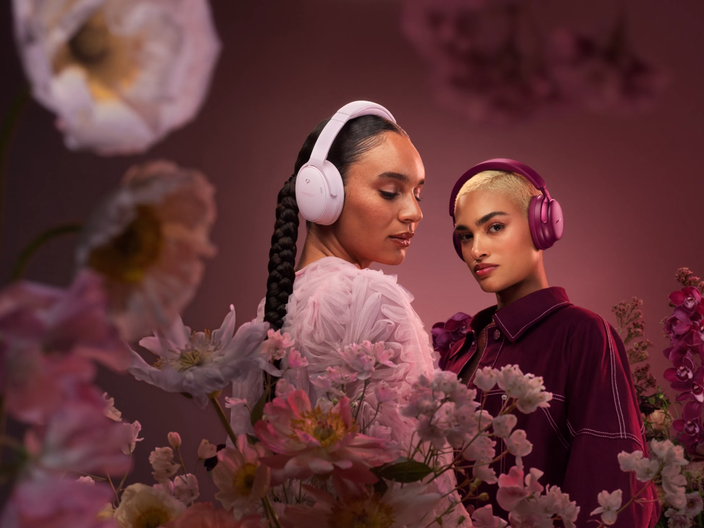 Two people wearing QuietComfort Ultra Headphones and QuietComfort Headphones in a pink environment with flowers around.