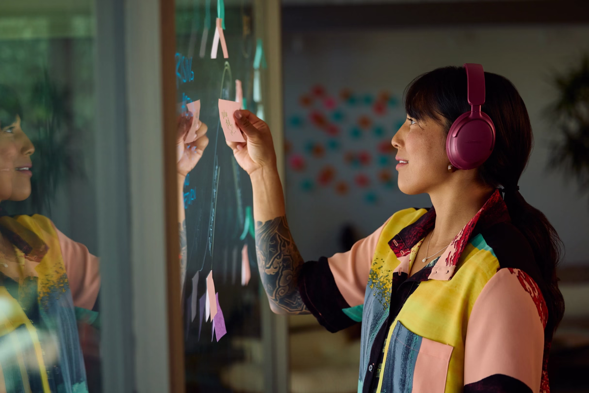 A woman wearing QuietComfort Ultra Headphones in Deep Plum putting sticky notes on a mirror covered in other sticky notes.